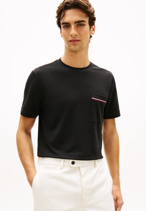 Ung mand iført en sort T-shirt med korte ærmer og en brystlomme med røde, hvide og blå striber, kombineret med hvide bukser.