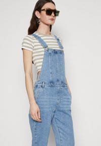 Pantalones de mezclilla en azul claro con tirantes ajustables, bolsillos frontales y un ajuste relajado. Combinados con una camiseta de manga corta a rayas.