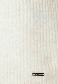 Strickpullover in Off-White, mit weicher Textur, geripptem Muster und einem kleinen metallischen Logo-Tag, das auf der Vorderseite angenäht ist.
