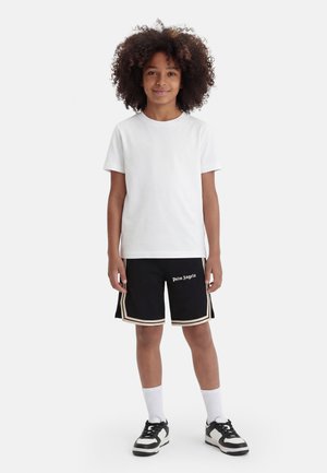 Barn med krøllet hår iført en hvid t-shirt, sorte "Palm Angels" shorts med beige kant, hvide sokker og sort-hvide sneakers, står stille.