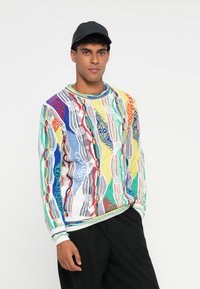 Πολύχρωμο πλεκτό πουλόβερ με γυαλιστερό μοτίβο σε μπλε, κίτρινο, κόκκινο και πράσινο. Στρογγυλή λαιμόκοψη και ριμπ μανσέτες. Φορεμένο με μαύρα παντελόνια.