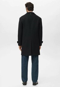 Abrigo de lana negro con corte recto, con una abertura en la parte trasera y puños abotonados. Lleva puesto sobre unos pantalones de mezclilla azul y zapatos.