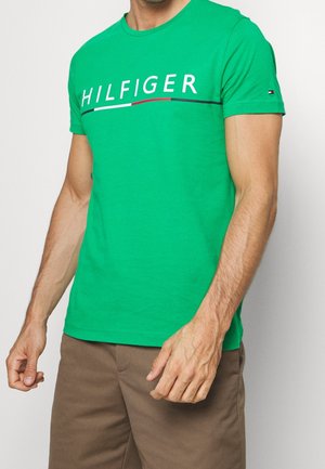 Homme portant un t-shirt vert vif à manches courtes avec le texte "HILFIGER" et des rayures de couleurs, associé à un pantalon marron.