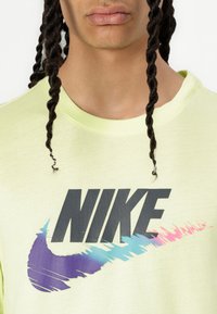Svijetložuta pamučna majica s velikim crnim Nike logotipom koji ima dizajn u obliku četkice s više boja u plavoj, ljubičastoj i ružičastoj.