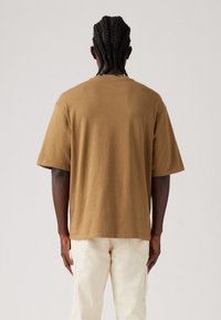 Tricou din bumbac maro, cu mânecă scurtă, cu un fit relaxat și guler standard, dintr-un material texturat, prezentat din spate pe un fundal neutru.