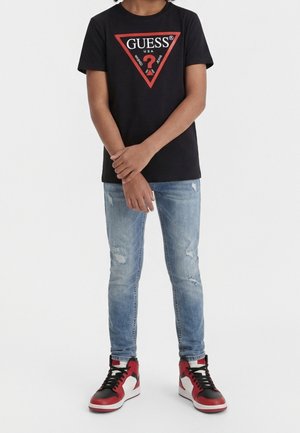 Person iført en sort Guess t-shirt med rød trekantslogo, lyseblå revnede jeans og røde, hvide og sorte høje sneakers, står med hænderne foldet.