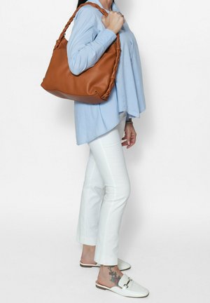 Borsa a spalla in pelle tan con manico intrecciato, texture morbida e forma arrotondata. Indossata con una camicia blu chiaro e pantaloni bianchi.