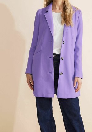 Femme portant un long manteau violet boutonné par-dessus un haut blanc et un jean bleu foncé sur un fond beige.