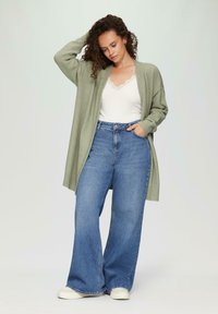 Helles grünes Strickcardigan, offene Vorderseite, gerippte Ärmelbündchen, getragen über einem weißen Oberteil mit Spitzenbesatz. Kombiniert mit hochtaillierten, weit geschnittenen blauen Jeans und weißen Turnschuhen.