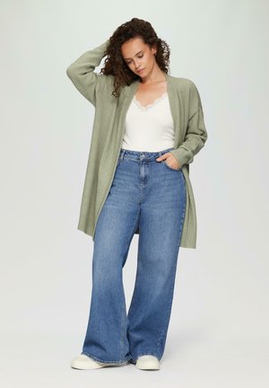 Cardigan tricoté vert clair, à l'avant ouvert, avec des poignets côtelés, porté sur un haut avec des dentelles blanches. Assorti à un jean bleu taille haute, à jambes larges et des baskets blanches.