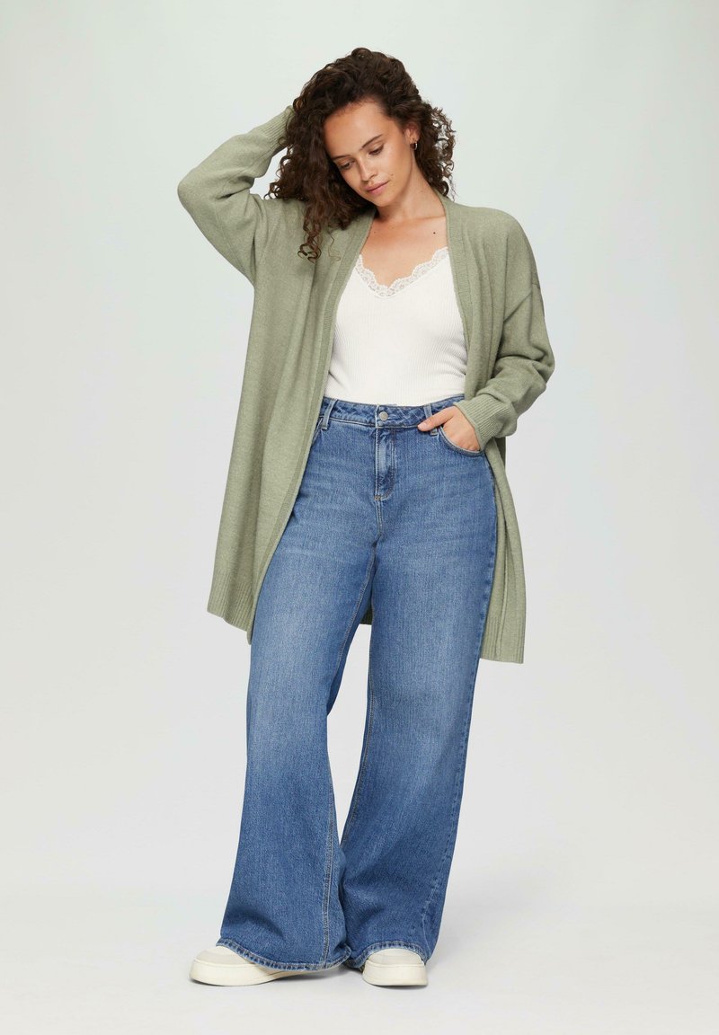 Helles grünes Strickcardigan, offene Vorderseite, gerippte Ärmelbündchen, getragen über einem weißen Oberteil mit Spitzenbesatz. Kombiniert mit hochtaillierten, weit geschnittenen blauen Jeans und weißen Turnschuhen.