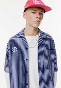 Korte mouwen gestreept shirt in blauw en wit katoen met een zak en geborduurd logo, gecombineerd met een roze pet. Zilveren ketting zichtbaar.