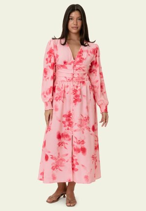 Frau trägt ein rosa Midikleid mit langen Ärmeln und rotem Blumenmuster, Knopfleiste vorne, tailliert und mit gerafften Bündchen, steht mit neutralem Gesichtsausdruck.