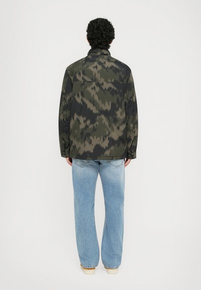 Kamuflage-mønsteret jakke med høj krave, lavet af et let stof; parret med falmede blå vide jeans og beige sko.