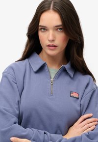 Sudadera de forro polar color púrpura claro con collar con cremallera, que presenta un parche rectangular con una bandera y un logotipo. Textura suave y ajuste relajado.