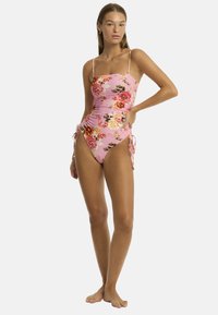 Un mannequin pose dans un maillot de bain une pièce à fleurs roses avec des liens latéraux réglables, affichant un look prêt pour la plage sur un fond uni.