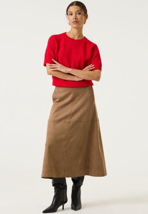 Eine Frau steht mit verschränkten Armen, trägt einen roten kurzärmeligen Pullover, einen beigefarbenen A-Linien-Rock und schwarze Stiefeletten mit Absatz vor einem schlichten Hintergrund.