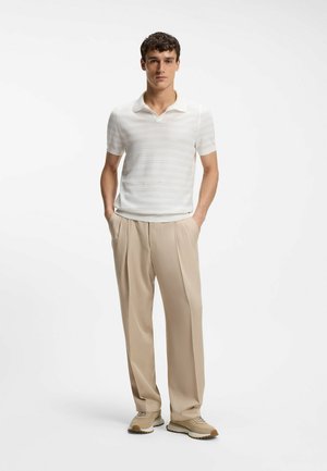 Young man standing with hands in pockets wearing white striped short-sleeve polo, beige pleated trousers, and beige sneakers.