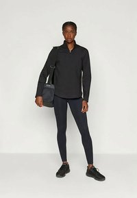 Fekete sportöltözetbe öltözött nő, aki egy válltáskával a vállán áll egy sima fehér háttér előtt.