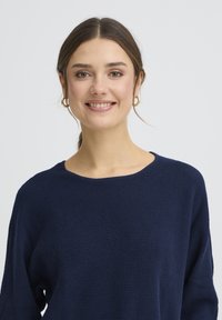 Pull en tricot bleu marine avec un large col et un motif texturé. Le tissu semble doux avec une coupe décontractée. Boucles d'oreilles dorées visibles.