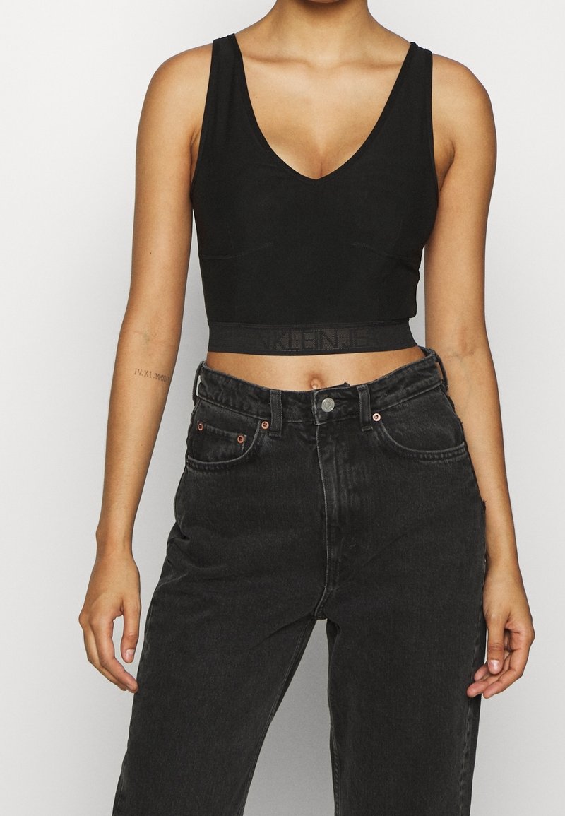 Débardeur noir court avec un décolleté en V et des bretelles larges, présentant une texture lisse et une bande élastique avec un détail de logo. Associe à un jean noir taille haute.