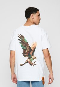 Hvid bomulds t-shirt med et stort, farverigt ørnegrafik på ryggen, med detaljerede fjer og et orange mærke ved kanten.