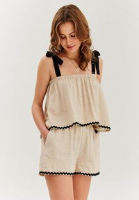 Béžové ľanové šaty s voľným vrchom, čiernymi stužkami a čiernym zúbkovaným lemom pozdĺž spodného okraja a šortiek; majú postranné vrecká.