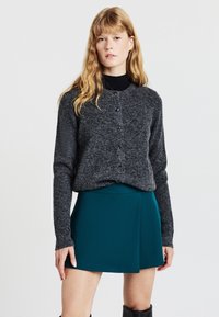 Giovane donna con lunghi capelli rossi che indossa un cardigan grigio scuro con bottoni sopra una maglietta nera, una gonna teal e stivali neri fino al ginocchio, in piedi.