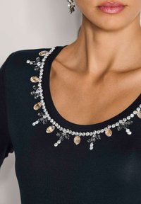 Close-up van de halslijn van een zwart top versierd met doorzichtige en amberkleurige druppelvormige rhinestones en decoratieve kralen.