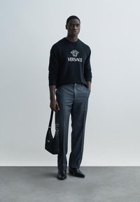 Μαύρο πλεκτό πουλόβερ με το λογότυπο "VERSACE", γκρι παντελόνι προσαρμοσμένης γραμμής, μαύρα παπούτσια και μαύρη τσάντα ώμου με χρυσές λεπτομέρειες. Ύφασμα με λεία υφή.