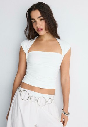 Ung kvinde med langt mørkt hår har en hvid ærmeløs top på, hvide bukser med stort sølvbælte med ring og sølvarmbånd på højre håndled.