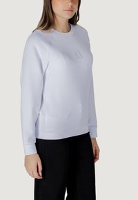 Sudadera azul clara con mangas raglán, logo bordado en el pecho, puños y dobladillo acanalados, combinada con pantalones negros.