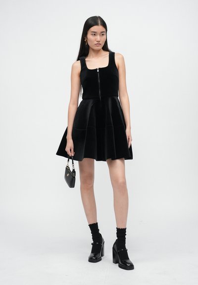 Femme portant une robe sans manches en velours noir avec fermeture éclair sur le devant, tenant un petit sac à main noir, assortie à des chaussettes noires et des talons blocs.