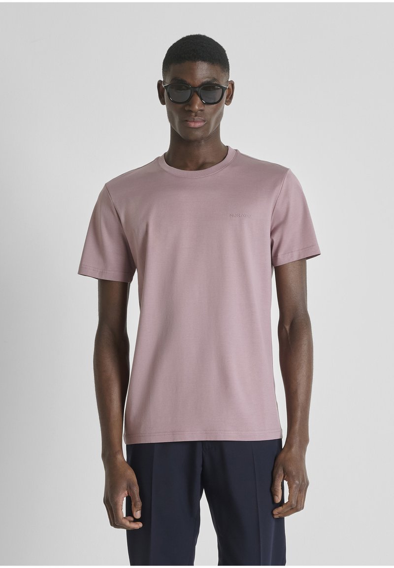 Hombre con gafas de sol oscuras, camiseta de manga corta color malva claro y pantalones azul marino oscuro, de pie frente a un fondo gris liso.