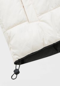 Kremowa kurtka puchowa z pikowanymi detalami, czarne wykończenie u dołu oraz regulowane zapięcie dla bezpiecznego dopasowania. Miękka, gładka tekstura.