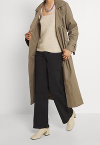 Canottiera beige in maglia sotto un trench coat khaki aperto, abbinata a pantaloni larghi neri e stivaletti a blocco color crema con tacco. Collana di catena in argento.