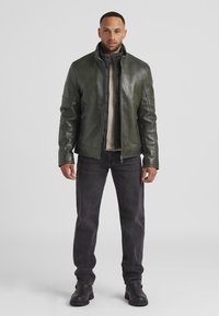 Dunkelgrüne Lederjacke mit hohem Kragen, Reißverschluss auf der Vorderseite und strukturierten Schulterpartien, kombiniert mit schwarzen Jeans und schwarzen Stiefeln.