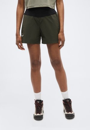 Άτομο που στέκεται, φοράει σκούρα πράσινα σορτς για υπαίθριες δραστηριότητες, μπεζ πεζοπορικά μποτάκια με κόκκινα κορδόνια, λευκές κάλτσες και ένα κοντό χακί τοπ.