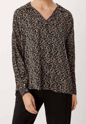 Femme portant un chemisier noir avec un motif géométrique abstrait beige et un pantalon noir, debout avec une main détendue le long du corps.