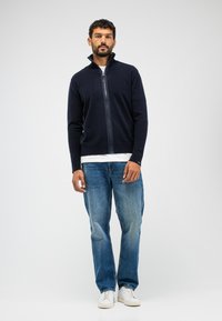 Marinefarbener Reißverschluss-Pullover aus Strickmaterial, mit hohem Kragen und gerippten Bündchen, kombiniert mit hellblauen Jeans und weißen Sneakers.
