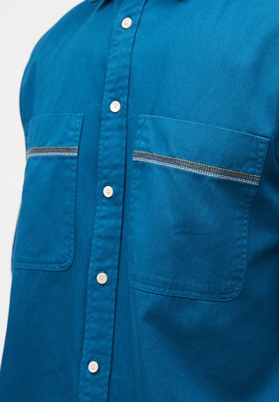 Πουκάμισο με κουμπιά σε τυρκουάζ χρώμα και δύο τσέπες στο στήθος. Διαθέτει ραφές σε αντίθετο χρώμα στις τσέπες και ύφασμα λείο με υφή.