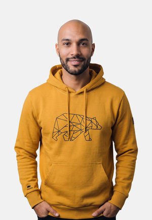 Glimlachende man met korte baard die een mosterdgele hoodie draagt met een geometrisch berenontwerp, staand met zijn handen in zijn zakken tegen een effen achtergrond.