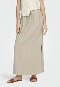 Falda maxi de lino beige con una abertura lateral y cintura elástica. Combinada con sandalias deslizantes de mimbre marrones. Tejido ligero y ajuste relajado.
