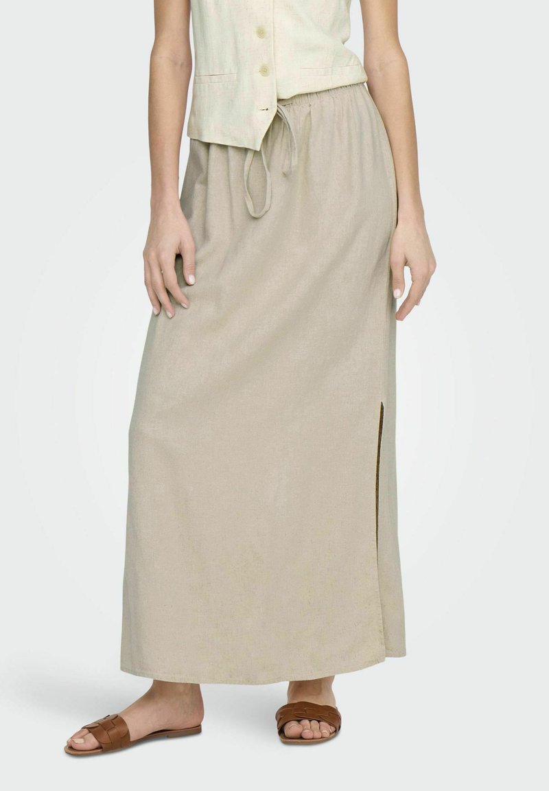 Falda maxi de lino beige con una abertura lateral y cintura elástica. Combinada con sandalias deslizantes de mimbre marrones. Tejido ligero y ajuste relajado.