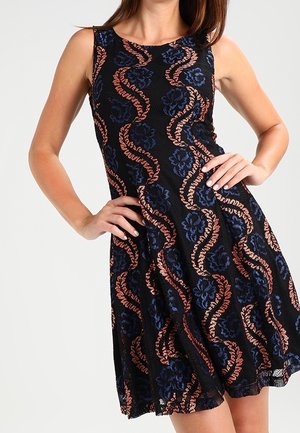 Robe noire sans manches avec des motifs floraux bleus et des tourbillons orange portée par une femme les mains sur les hanches.