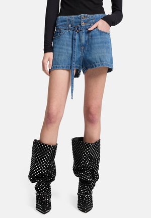 Kvinde iført blå denimshorts med bælte og slaskede sorte støvler med sølvnitter, står mod en ensfarvet baggrund.