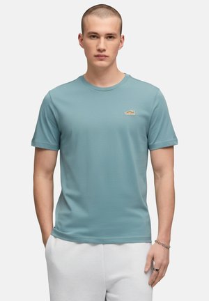 Giovane uomo con capelli corti che indossa una maglietta blu a girocollo e pantaloni bianchi, in piedi con le mani rilassate ai lati.