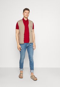Beige quiltet vest over en rød polo, kombinert med lyseblå oppbrettede jeans og grå sandaler. Enkelt design med funksjonelle lommer.