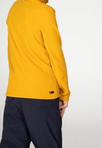 Žlutý fleecový svetr s vysokým límcem, texturovaná tkanina a černá štítková detaily na spodním lemu; spárováno s tmavě modrými kalhotami.