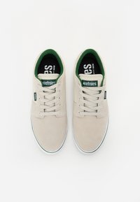 Beige suede sneakers with green accents, white rubber soles, and flat laces. Features a logo tag on the tongue and patterned insole.